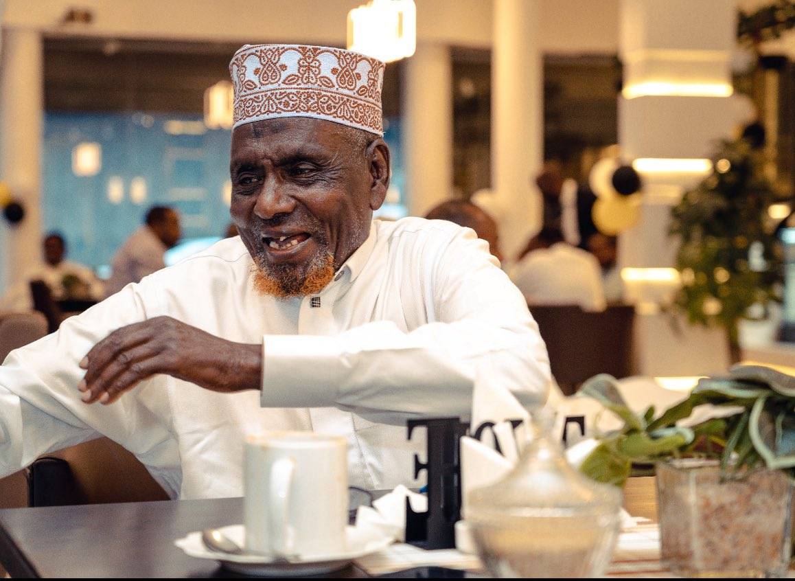 An older guest in a kofia seated at a restaurant table, mid-conversation